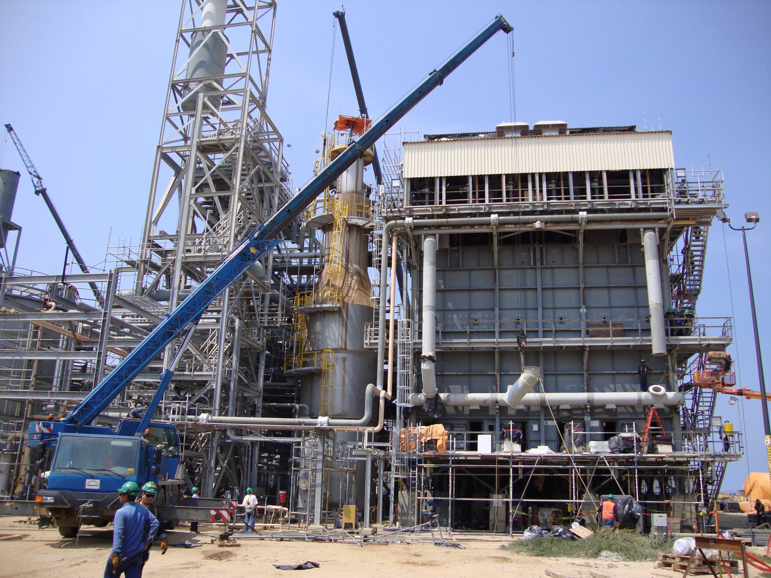 Imagen de una planta industrial en construcción con estructuras metálicas, tuberías y una grúa transportando material, y trabajadores con cascos de seguridad.