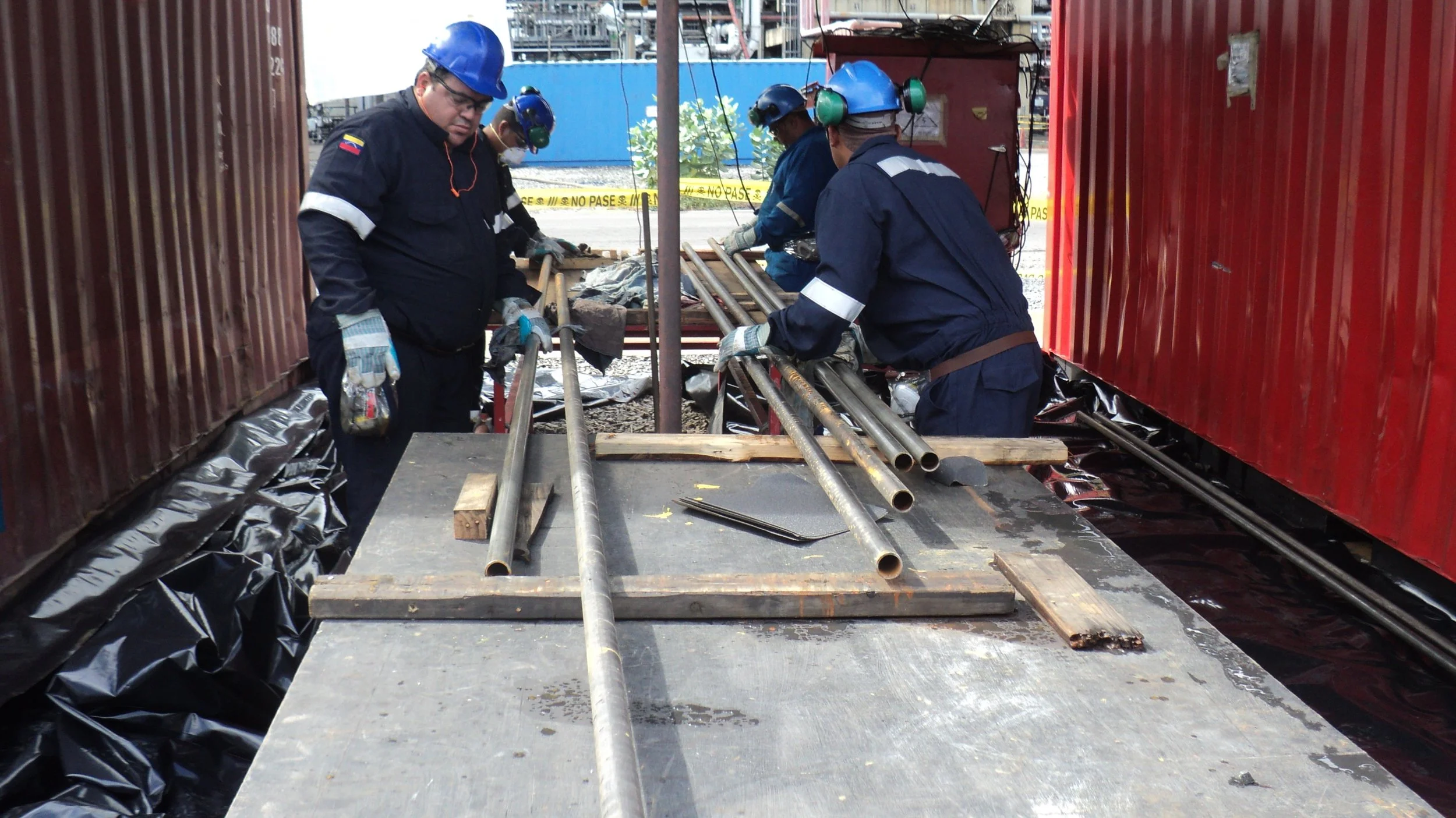 Trabajadores en un sitio de construcción, manipulando tuberías metálicas sobre una mesa de trabajo, con cascos y guantes de protección, entre contenedores de envío.