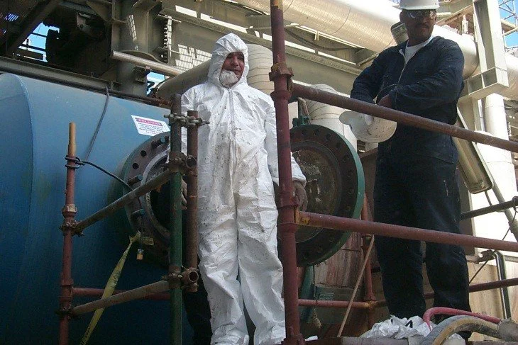 Dos hombres en un sitio industrial, uno con traje de protección blanco y otro con casco y uniforme oscuro, rodeados de tuberías y equipo industrial.