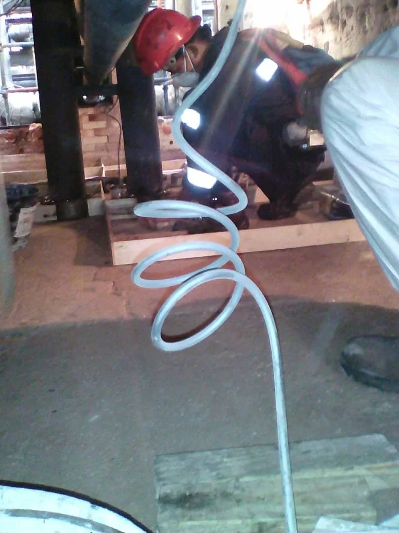 Persona con casco rojo y mascarilla, trabajando con herramientas en un entorno de construcción, mezclando o manipulando tubos de metal enrollados en espiral.