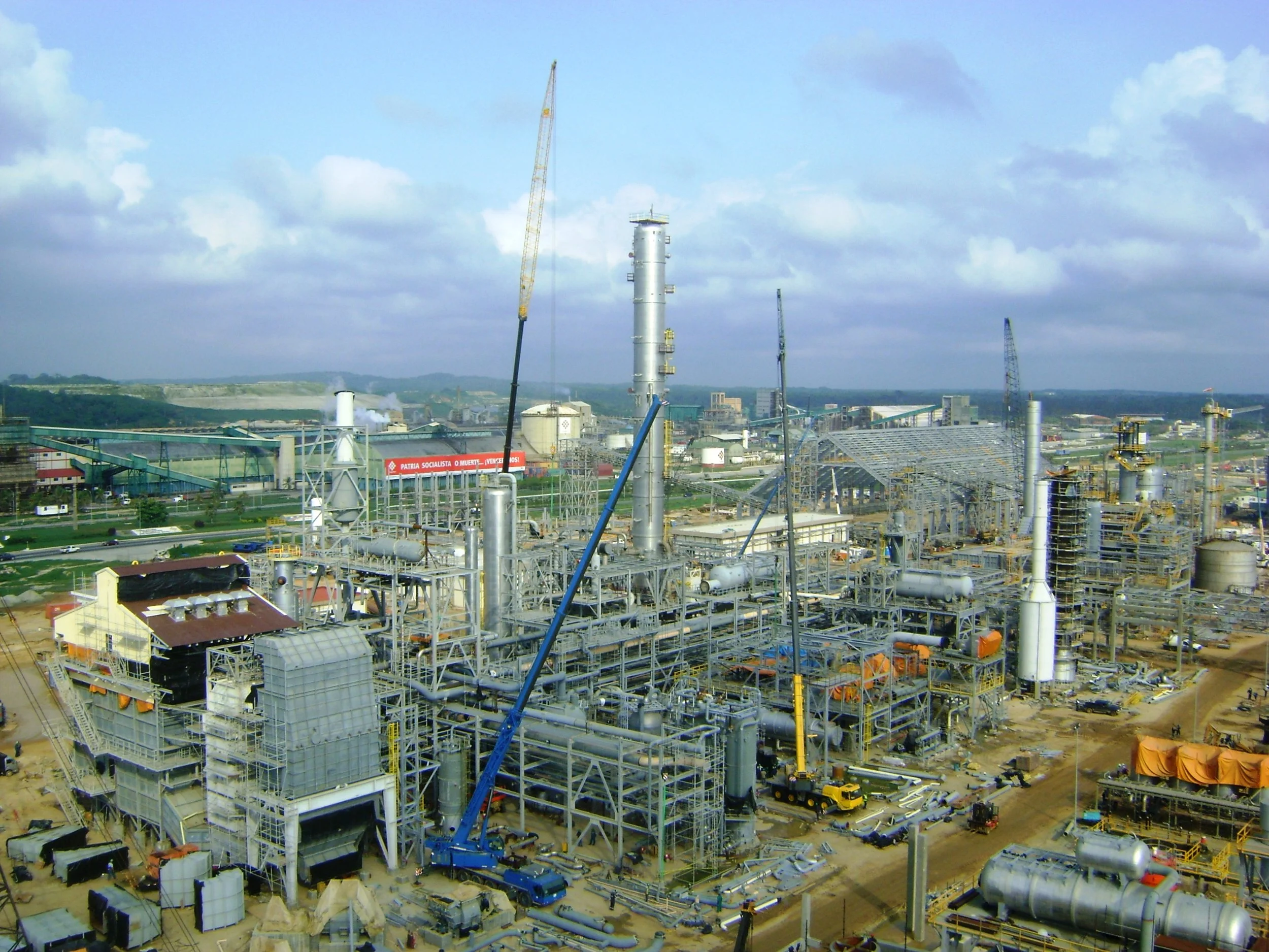 Planta Pequiven Amoniaco Moron Venezuela  con estructuras metálicas, torres y maquinaria en construcción, bajo cielo con nubes y área de tierra alrededor.