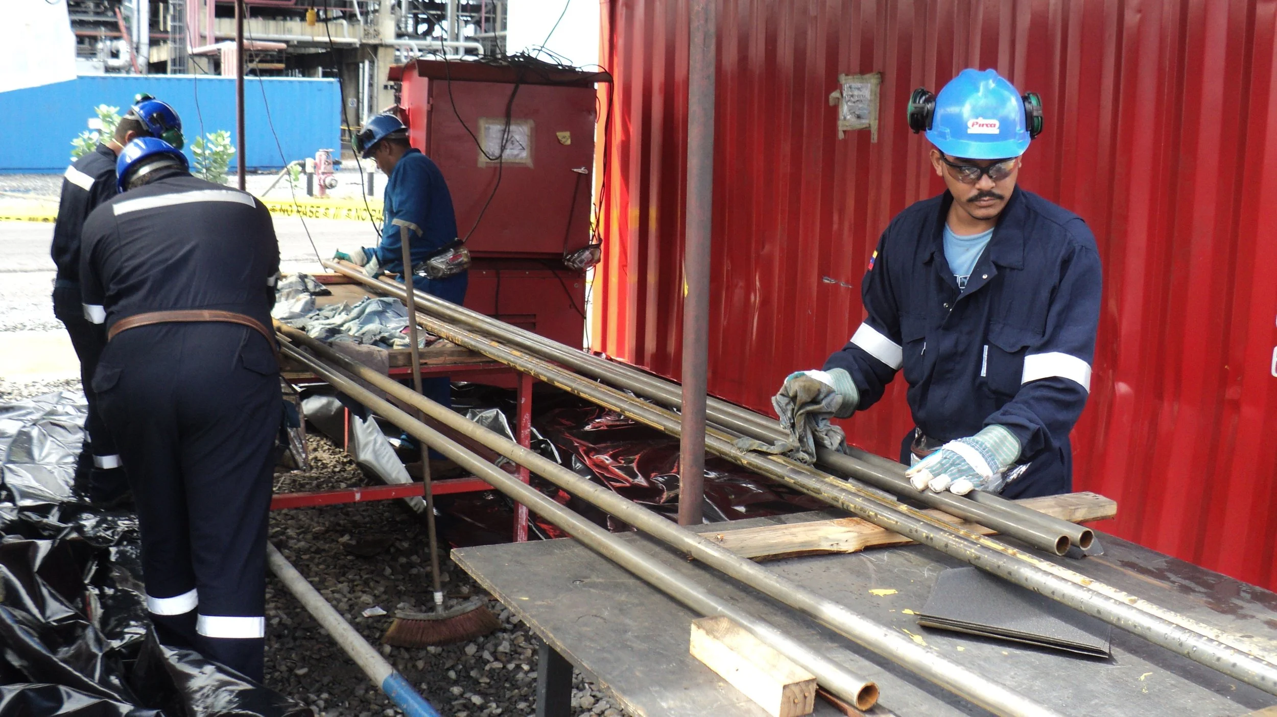 Trabajadores en equipo manipulando tuberías de metal en una obra con barreras de seguridad y equipo de protección, incluyendo cascos y guantes, en un entorno industrial o de construcción.