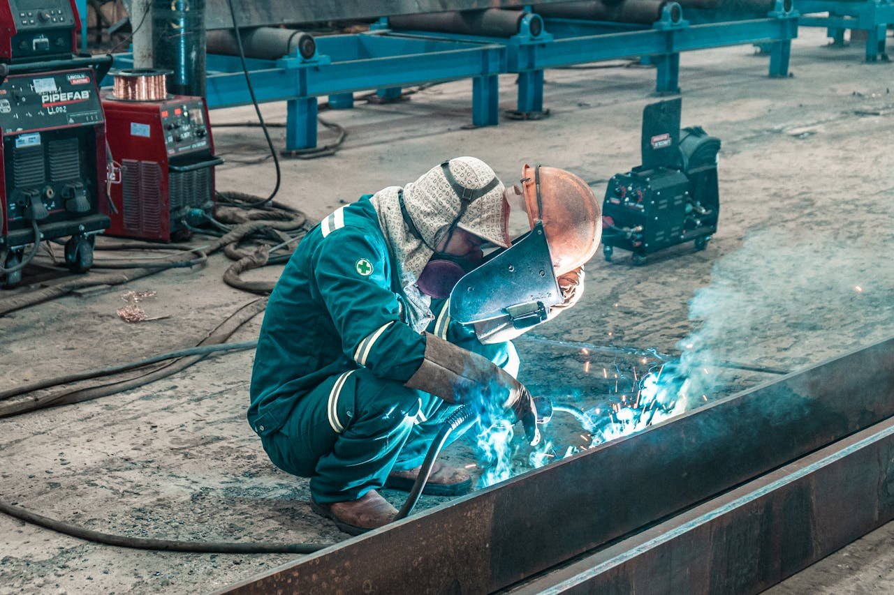 Trabajador soldando una estructura de metal en una fábrica, usando equipo de protección y máquinas de soldar en el fondo.