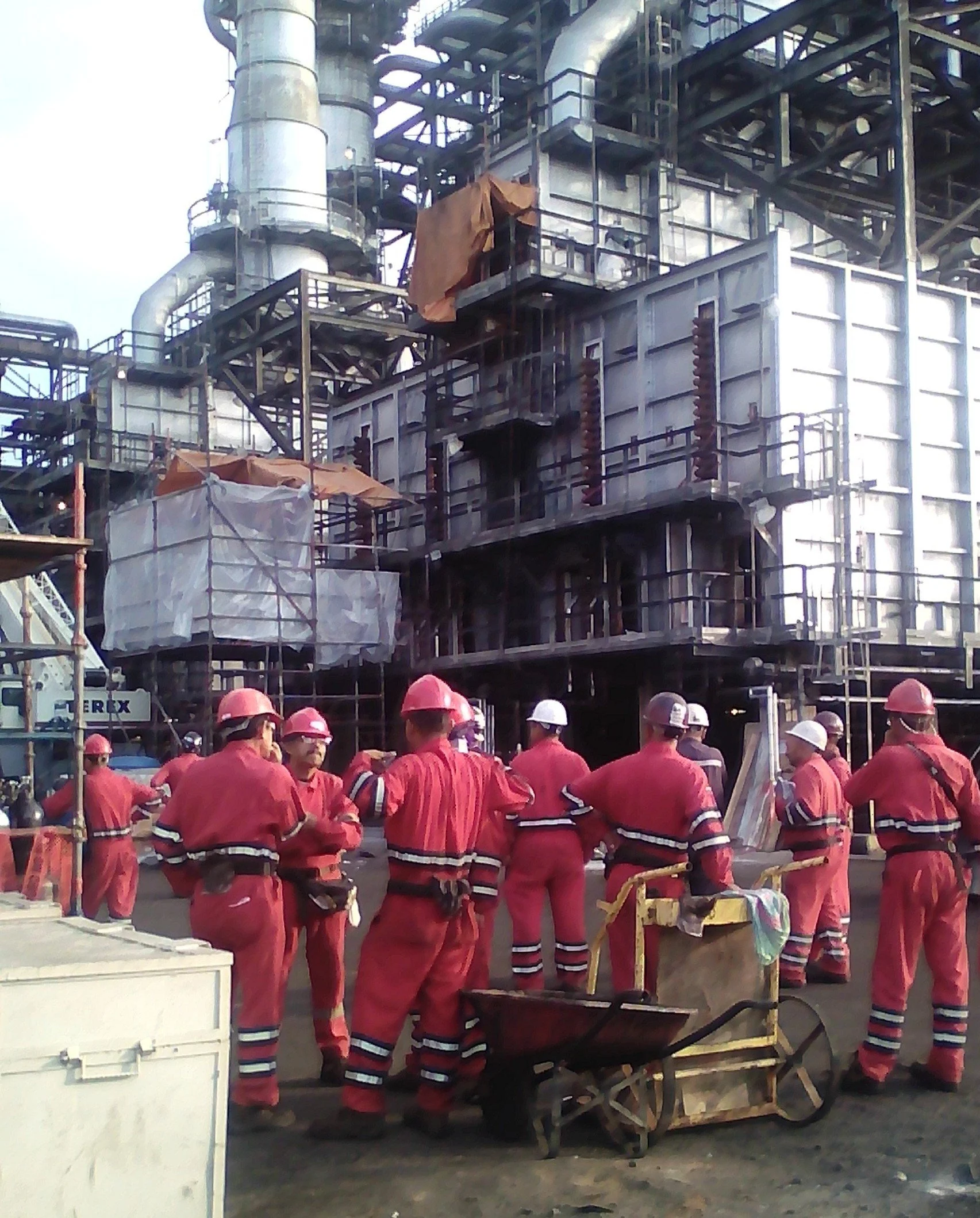 Grupo de trabajadores con equipo de protección, incluyendo cascos, en una planta industrial , con estructuras metálicas y tuberías en el fondo.