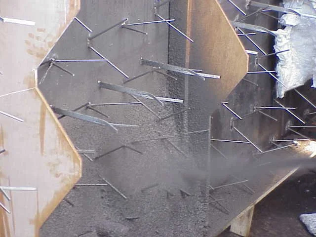 Dos trabajadores en equipo usando casco, gafas y máscara de protección, trabajando en una Estructura de acero con anclajes refractarios en un sitio de construcción.