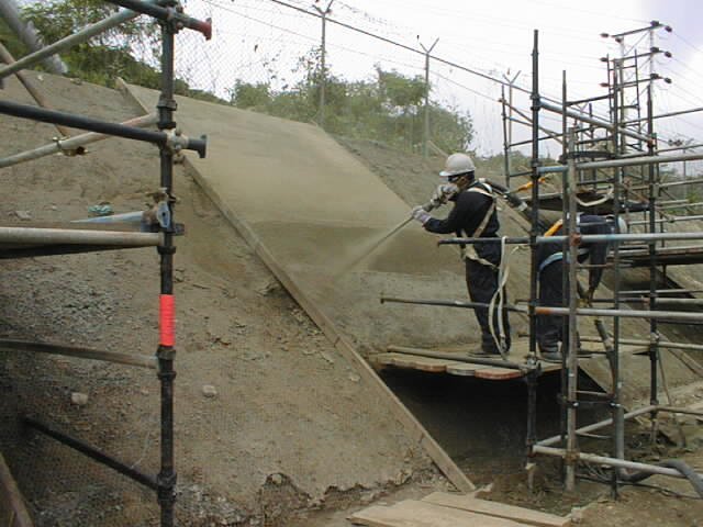 Trabajadores en una obra en construcción, proyectando cemento en una pendiente sobre una estructura de andamios.
