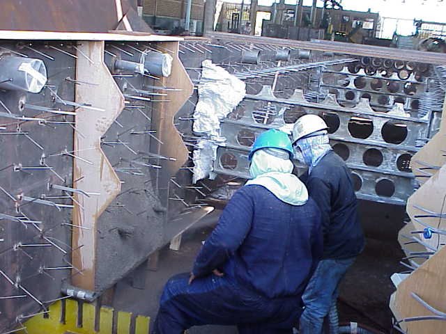 Dos trabajadores en equipo usando casco, gafas y máscara de protección, trabajando en una Estructura de acero con anclajes refractarios en un sitio de construcción.