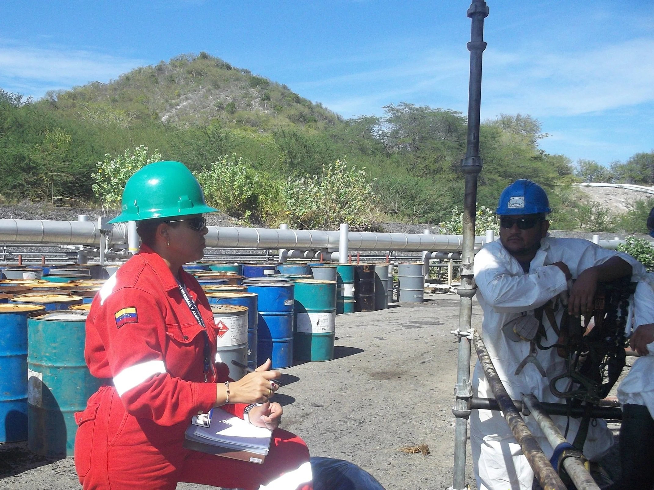 Dos trabajadores en una planta industrial, una mujer con casco verde y uniforme rojo, con gafas y anotando en un cuaderno, y un hombre con casco azul y ropa blanca, apoyado en estructura de metal, en un entorno con barriles y vegetación al fondo.