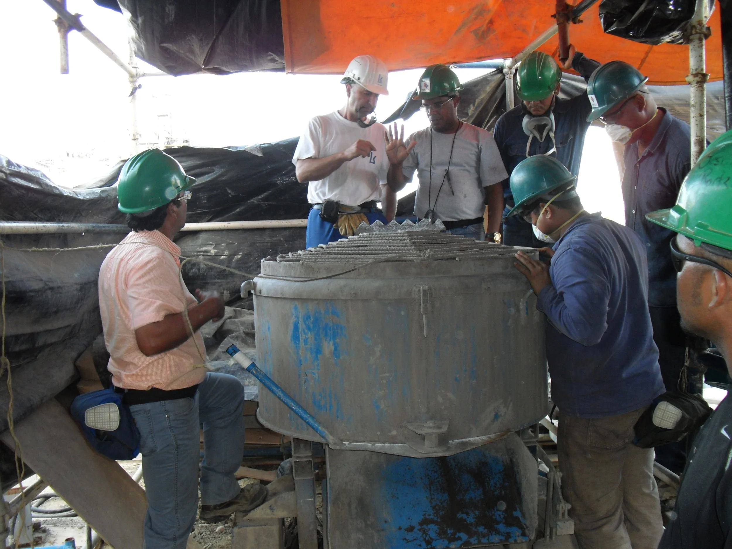 Varias personas con cascos verdes y ropa de trabajo observando y discutiendo encima de una máquina grande en construcción, en un sitio con estructura de andamios y toldos.