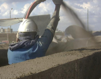 Un trabajador usando casco y ropa de protección, proyecta concreto en un sitio de construcción al aire libre.