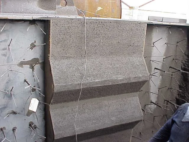 Dos trabajadores en equipo usando casco, gafas y máscara de protección, trabajando en una Estructura de acero con anclajes refractarios en un sitio de construcción.