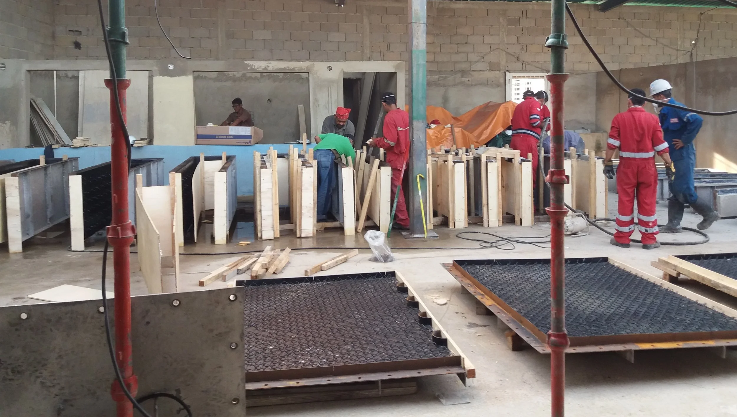 Trabajadores de construcción en un sitio en interiores, colocando moldes de madera para fundición o encofrado, con equipos y estructuras metálicas y madera alrededor.