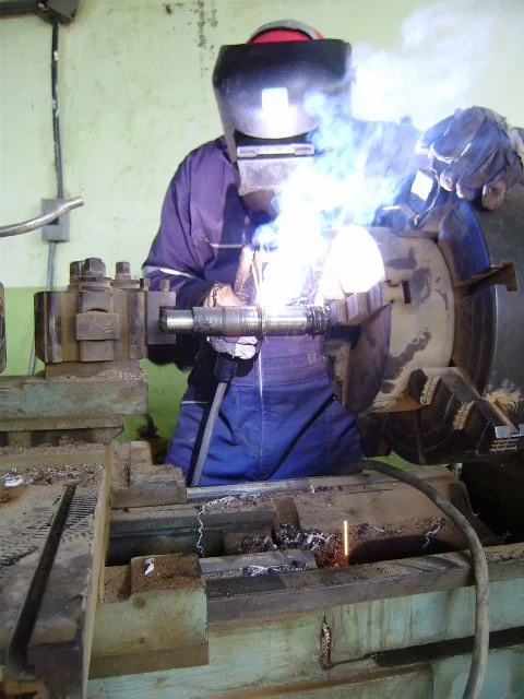Operario soldando una pieza metálica en un entorno de taller, con humo y luz brillante de la soldadura.