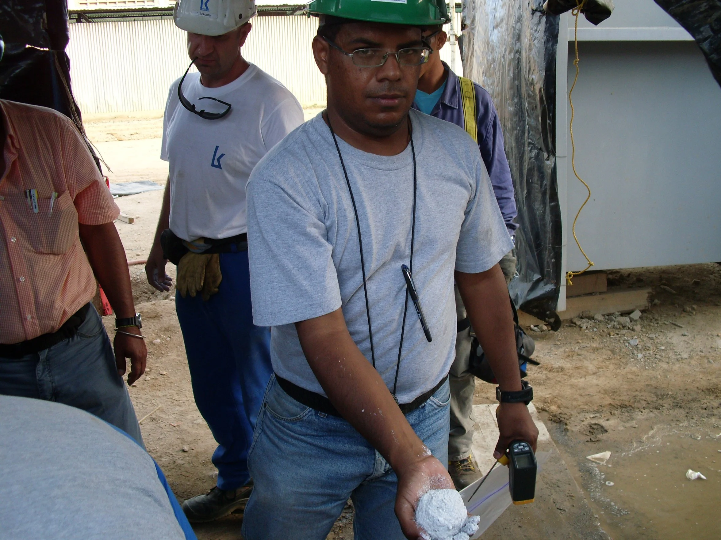 Hombre con casco verde, gafas y camiseta gris sostiene un instrumento en un sitio de construcción, rodeado de otros hombres.