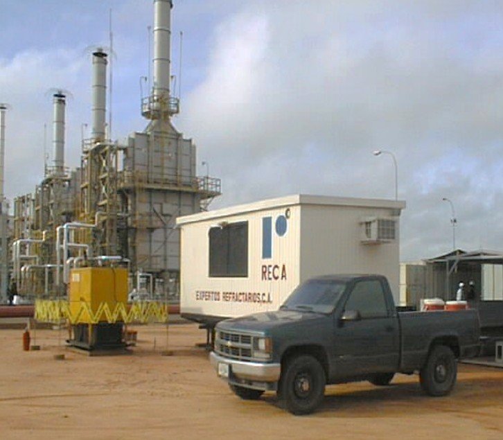 Planta de proceso industrial petrolera con maquinaria y un camión negro, carteles de RECA y letras en un contenedor.