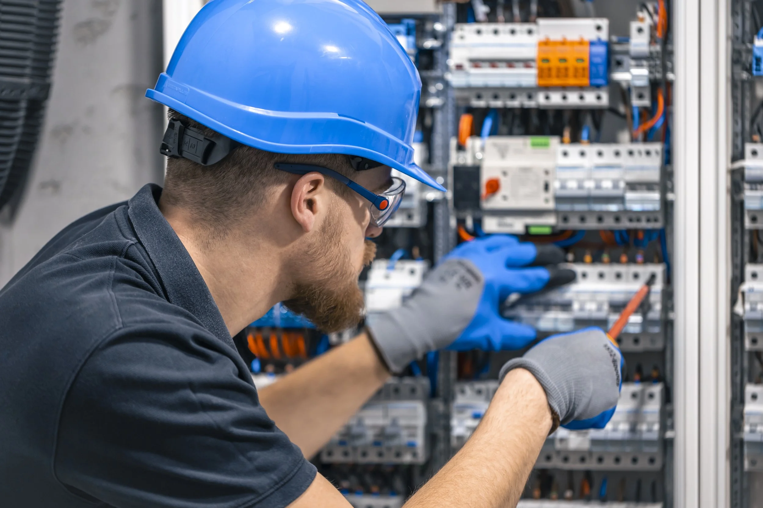 Un técnico con casco azul y guantes trabaja en un panel eléctrico con componentes y cables.