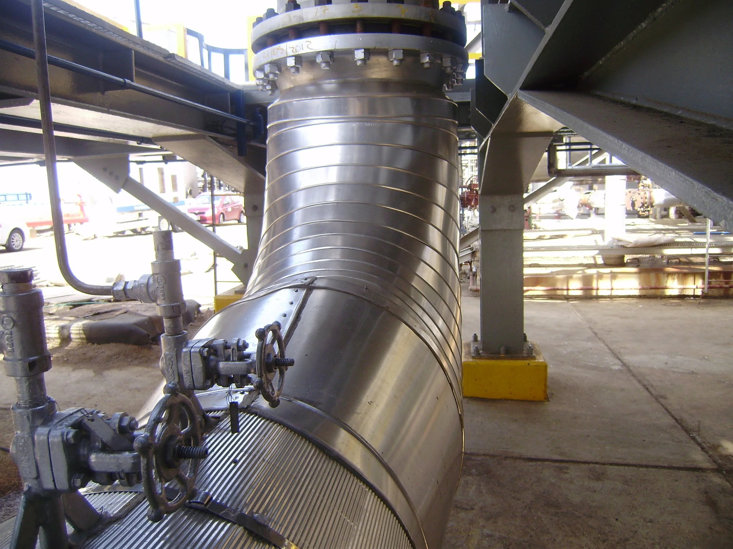 Tubería metálica grande en una construcción industrial, con válvulas y soporte, en un lugar abierto con vehículos en el fondo.