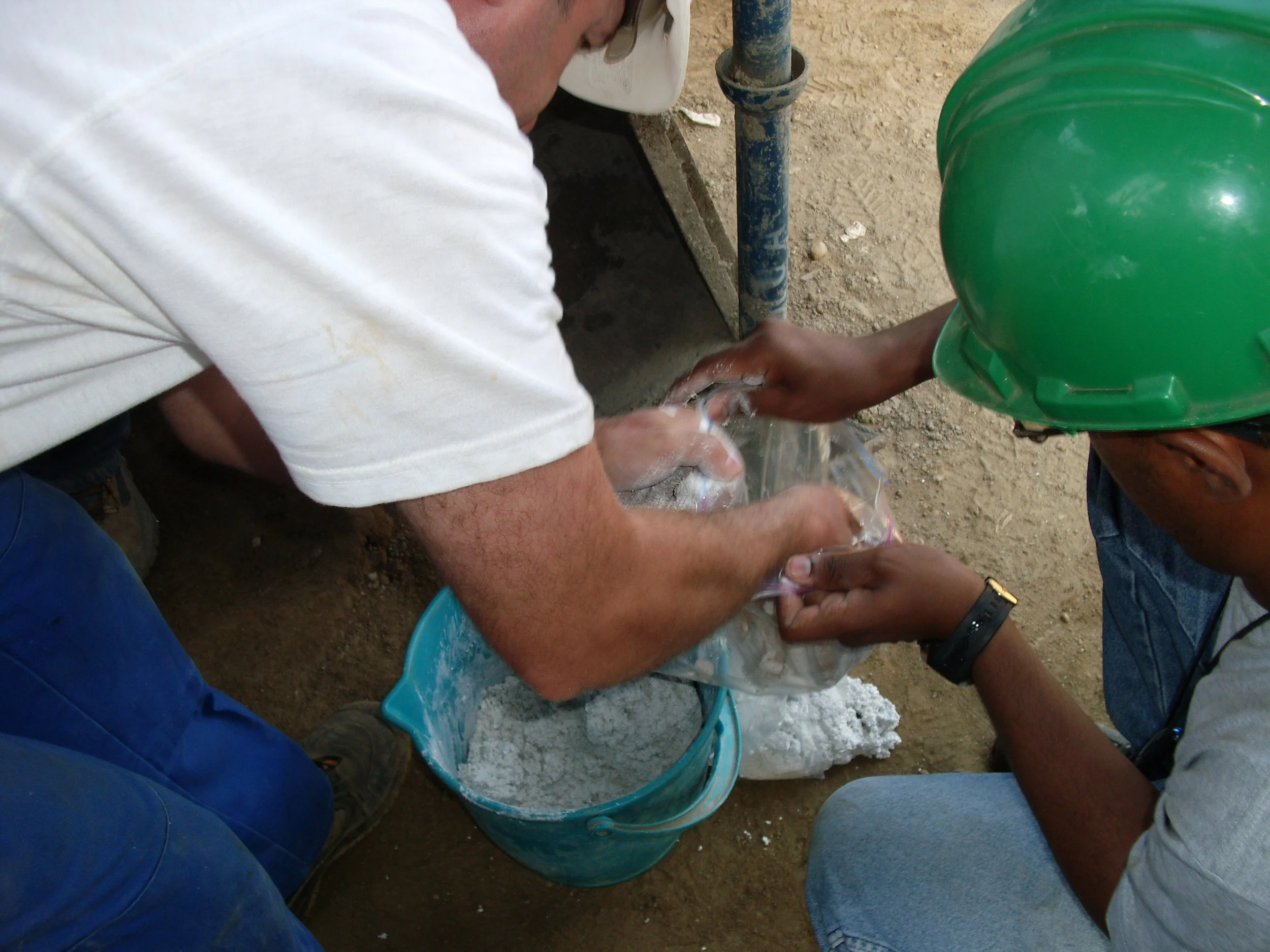 Dos hombres trabajando en la construcción, uno con casco verde y el otro sin casco, mezclando cemento en una bolsa de plástico en el suelo.