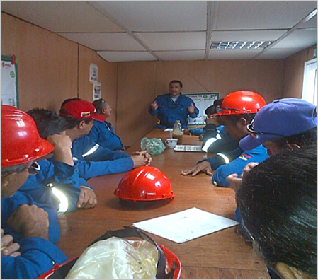 Grupo de trabajadores con cascos de seguridad en una sala de capacitación, escuchando a un instructor.