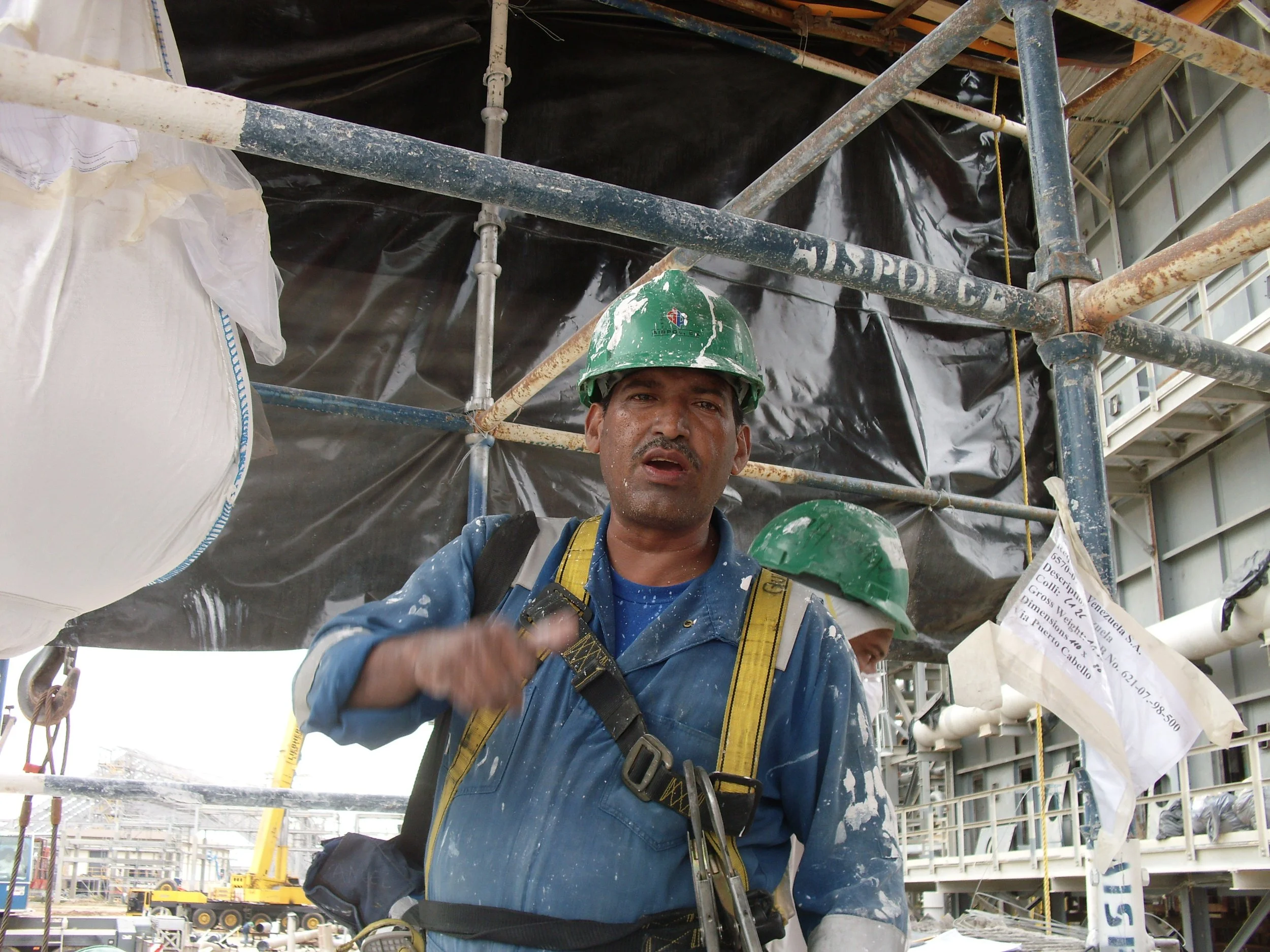 Obrero en casco verde en un sitio de construcción, con estructura metálica y maquinaria en el fondo.