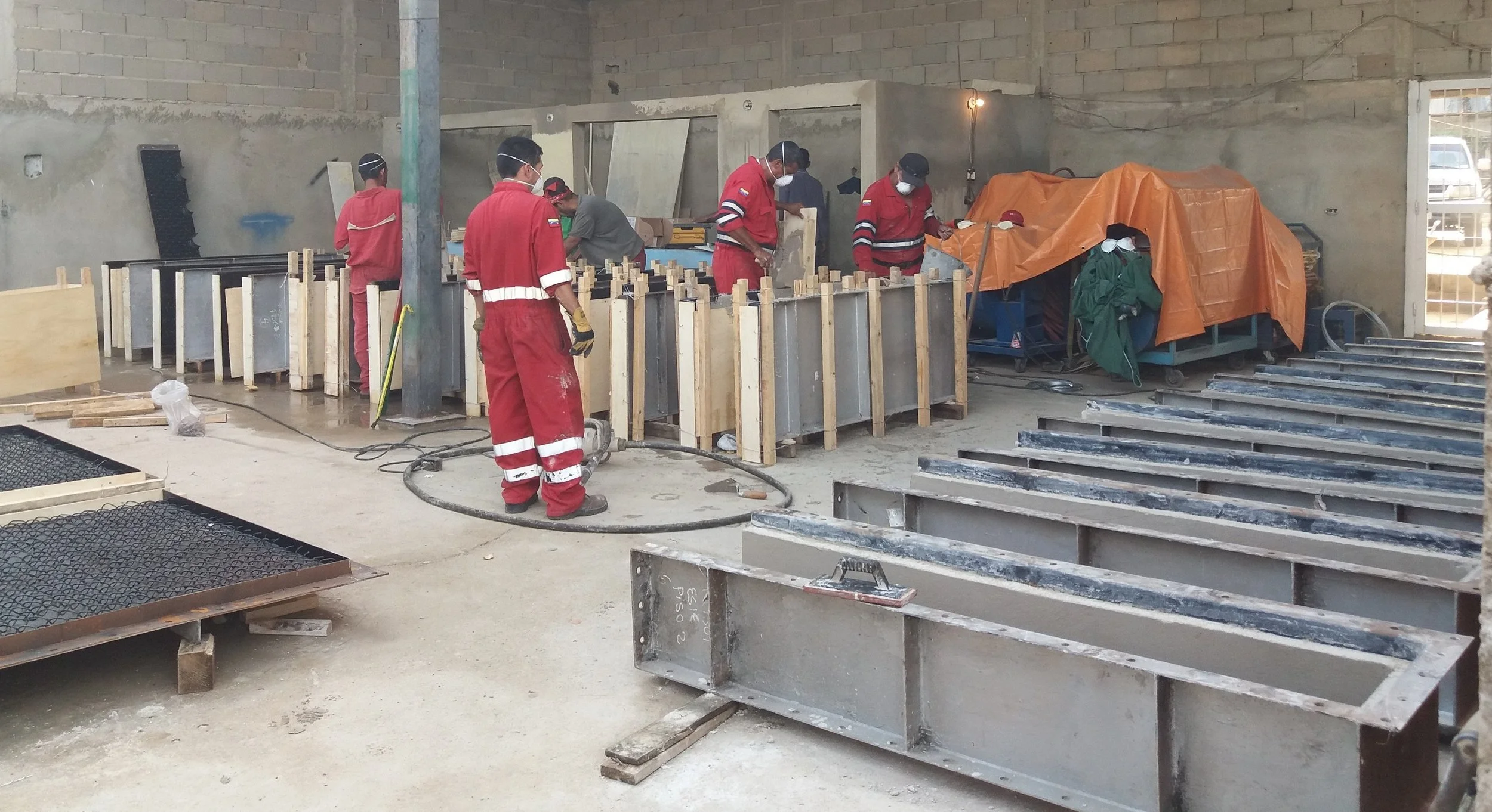 Trabajadores en una construcción, algunos con ropa de protección roja, otros con próxima a verde, algunos usando mascarillas y guantes. Hay estructuras metálicas y madera en el sitio de construcción.