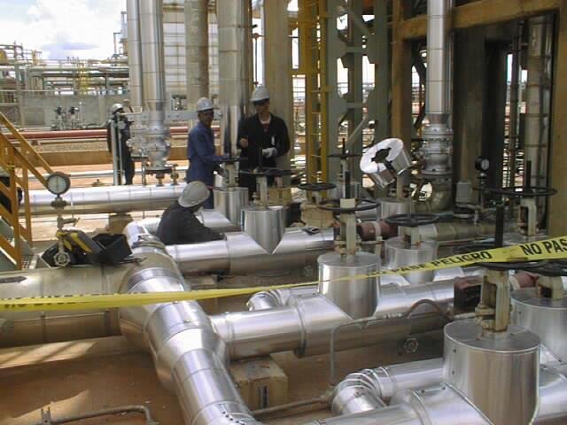 Trabajadores en una planta industrial revisando tuberías y maquinaria. El área está restringida con cinta amarilla de advertencia.