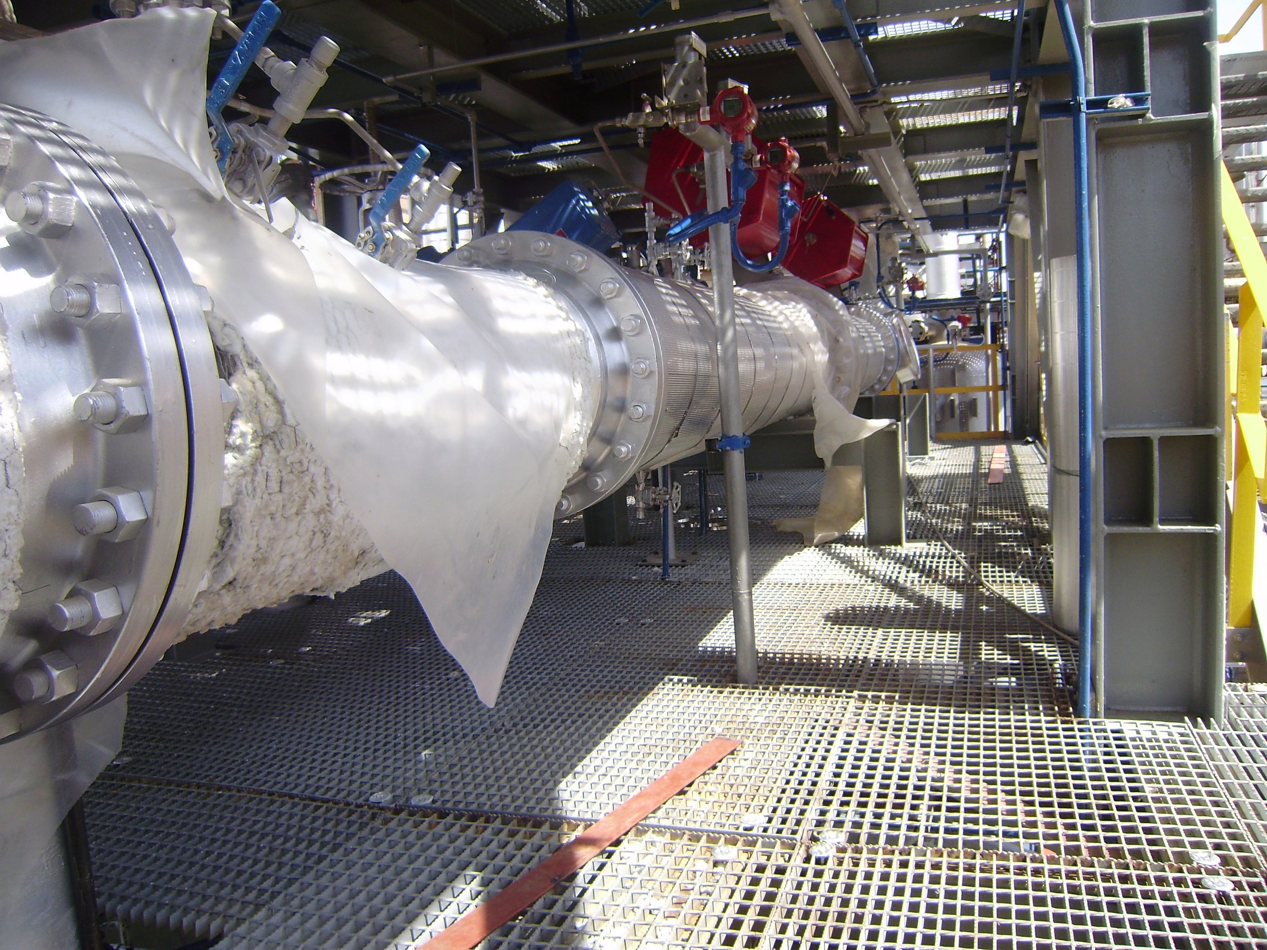 Vista de una tubería metálica grande con varias válvulas y conexiones en una estructura industrial, con una rejilla metálica en el piso y andamios alrededor.