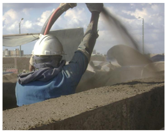 Un trabajador usando casco y ropa de protección, proyecta concreto en un sitio de construcción al aire libre.