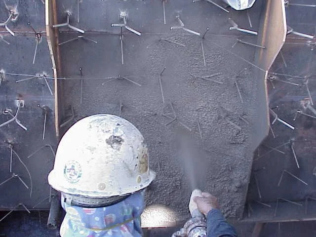 Dos trabajadores en equipo usando casco, gafas y máscara de protección, trabajando en una Estructura de acero con anclajes refractarios en un sitio de construcción.