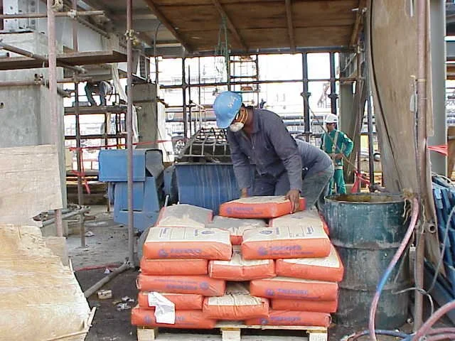 Trabajador en construcción con casco azul y máscara facial, colocando bolsas de cemento sobre un pallet en un sitio de obra.