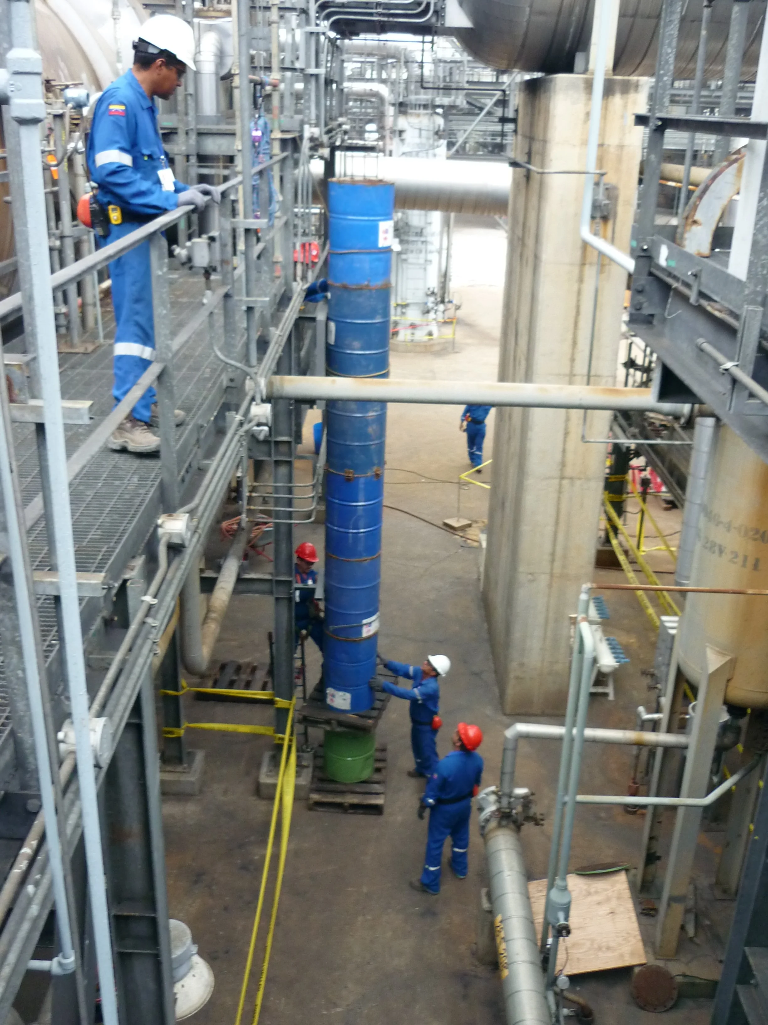 Trabajadores en un entorno industrial, algunos con cascos rojos y blancos, manipulando un tubo azul grande en medio de una estructura metálica.