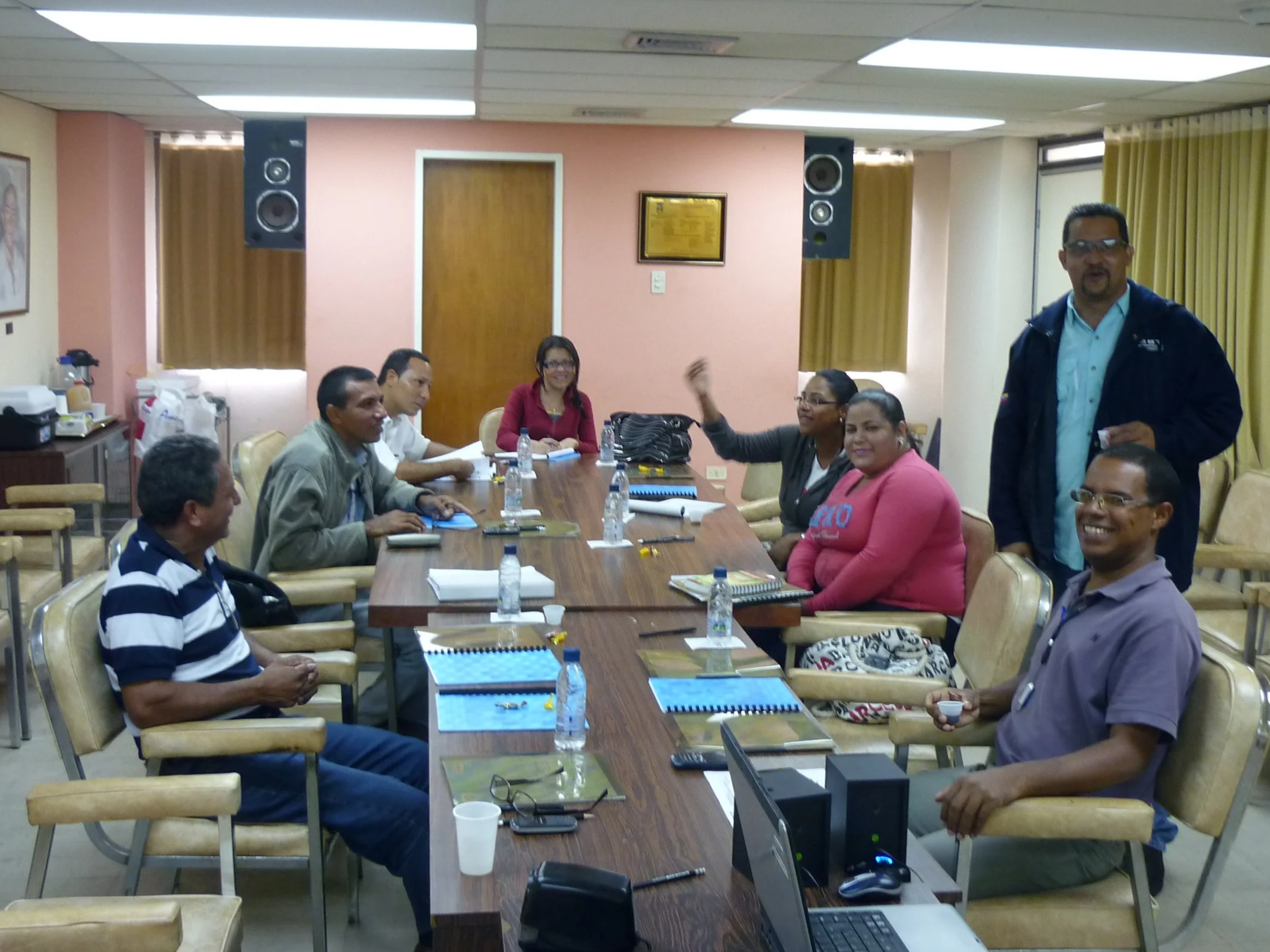 Reunión de varias personas en una sala de juntas, algunas sentadas y otras de pie, con mesas, laptops, documentos y botellas de agua sobre la mesa.