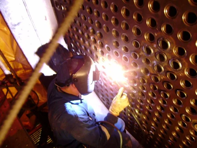 Trabajador soldando en un espacio cerrado, usando equipo de protección frente a una pared metálica perforada.