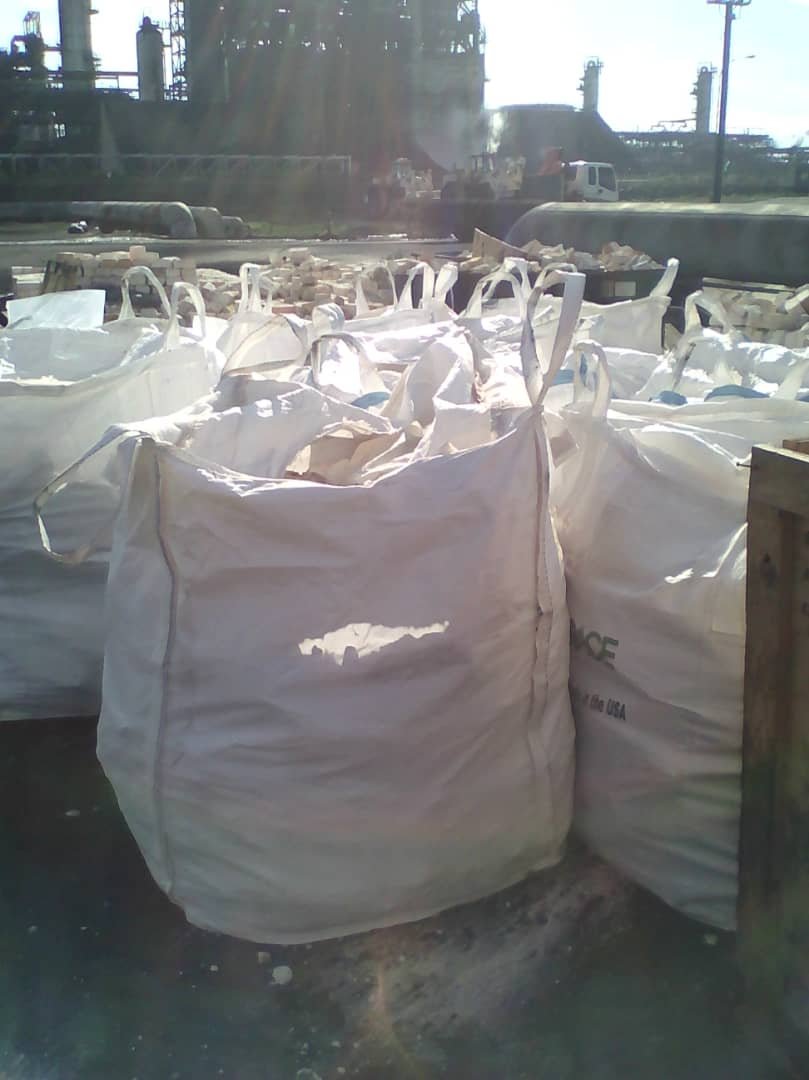 Grandes sacos blancos de plástico en un área industrial, rodeados de escombros y maquinaria, con estructura de fábricas al fondo.