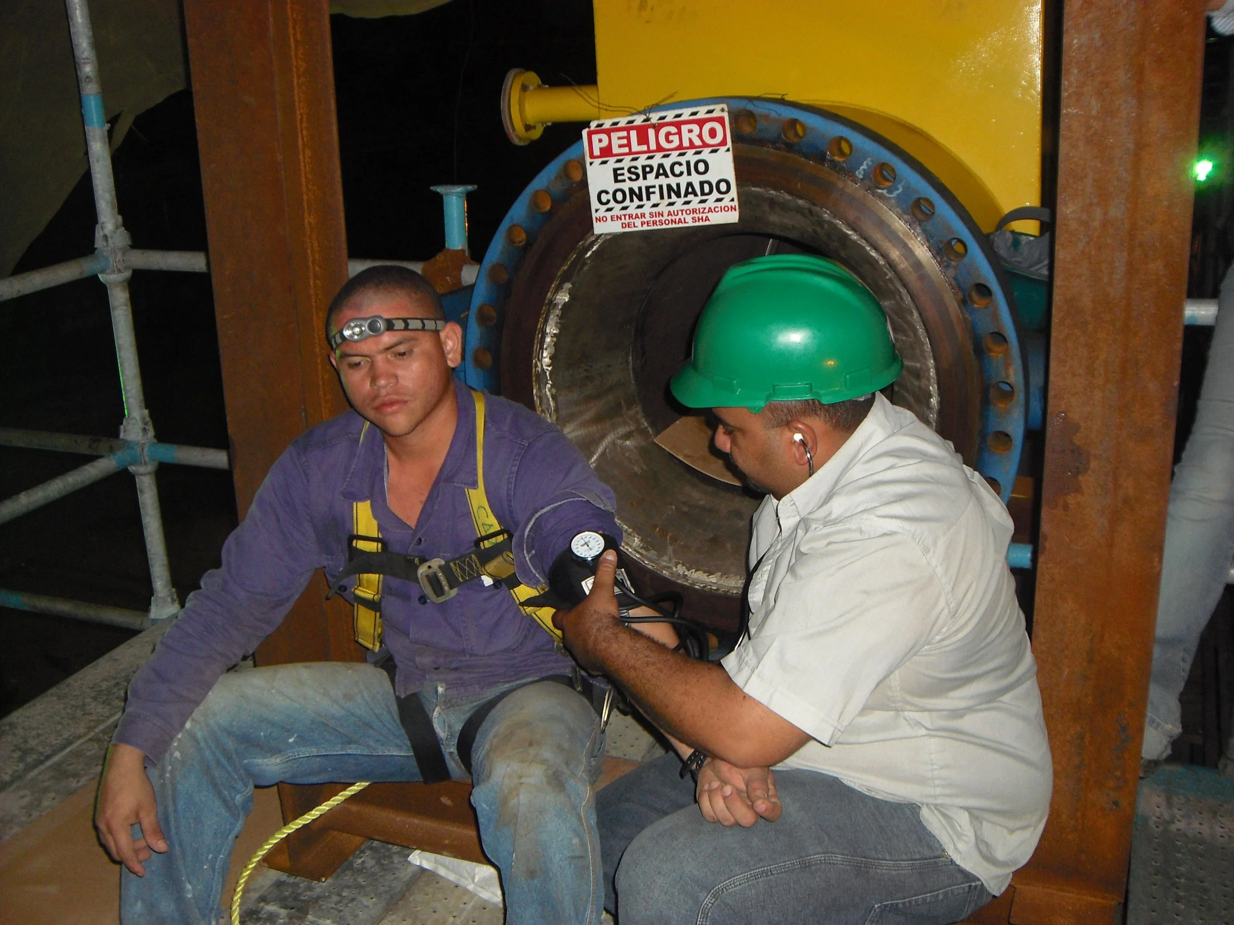 Dos trabajadores en equipo, uno con casco verde y escucha en los oídos, el otro con linterna en la cabeza, trabajando en una gran tubería industrial en un entorno de construcción.