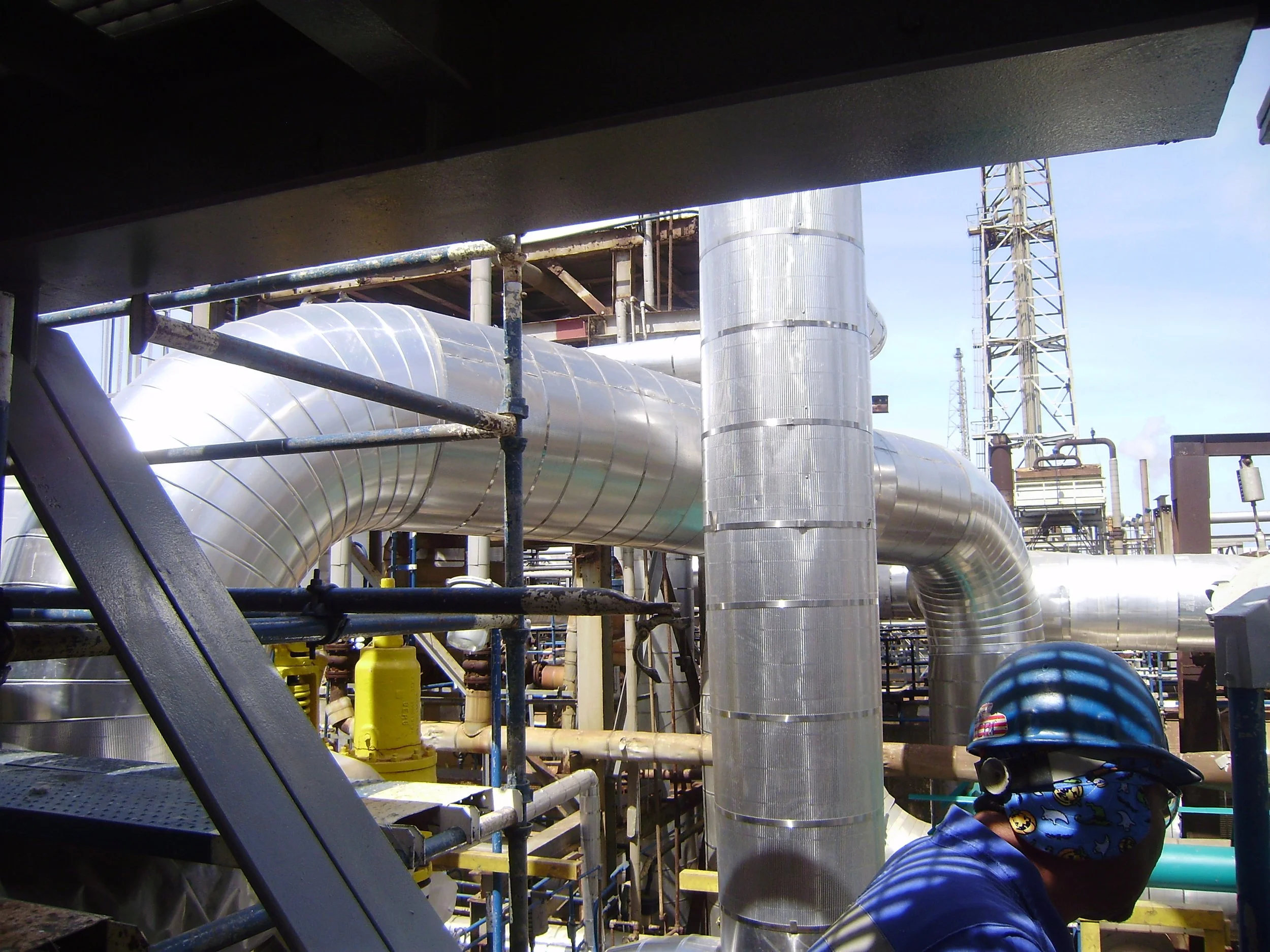 Una persona con casco azul y máscara en una refinería de petróleo, rodeada de tuberías y estructuras industriales de metal.