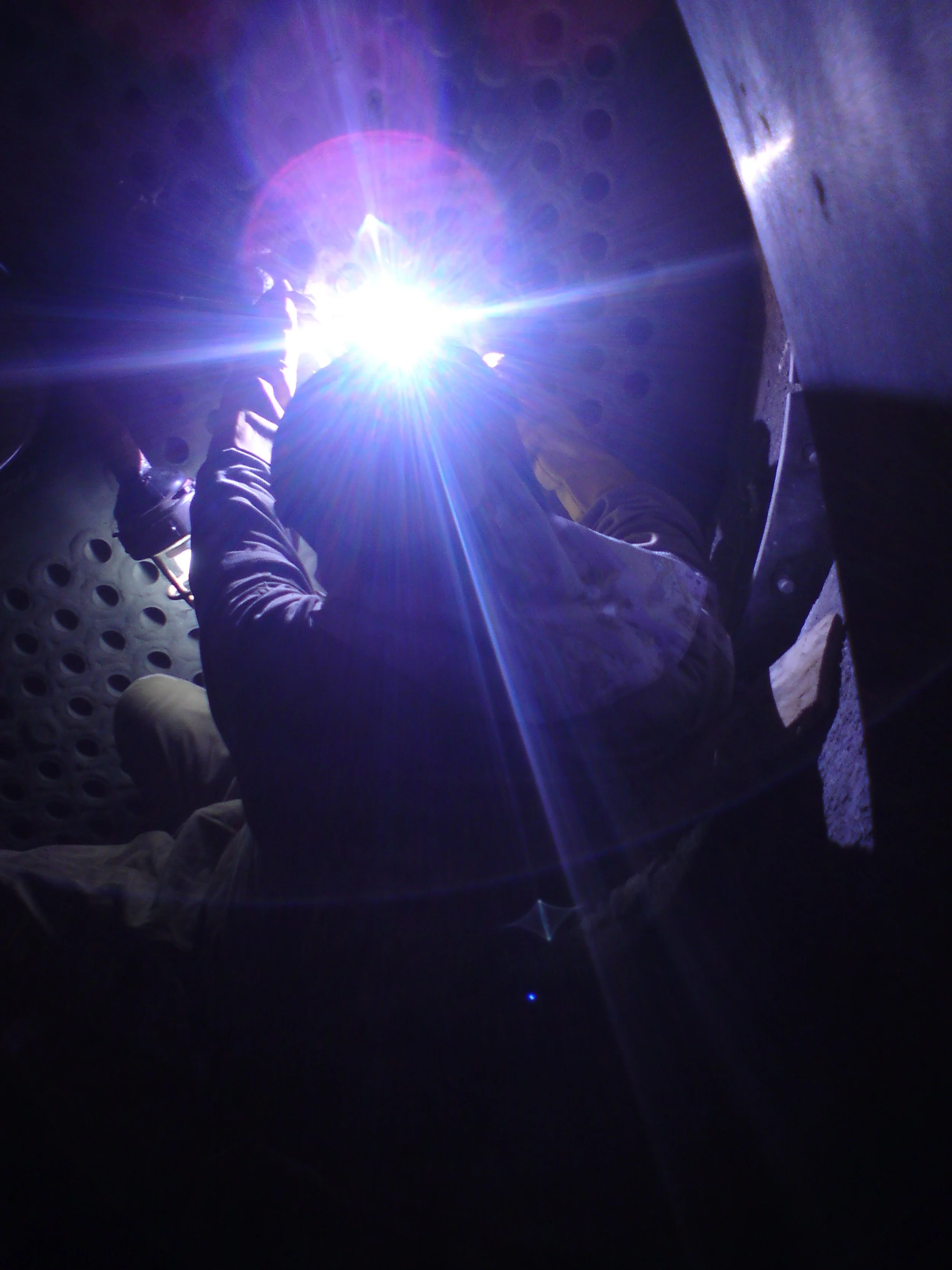 Un trabajador realiza soldadura en un entorno oscuro, con luz brillante proveniente de la soldadura que ilumina su rostro y manos.