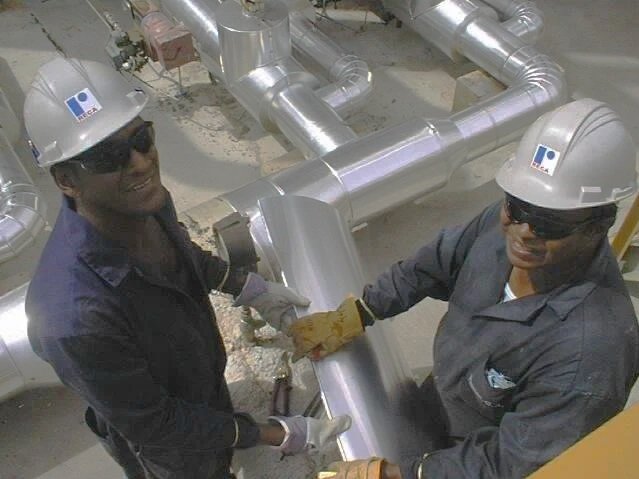 Dos trabajadores en casco y protección, estrechan sus manos sobre una tubería industrial de acero en una planta de construcción.