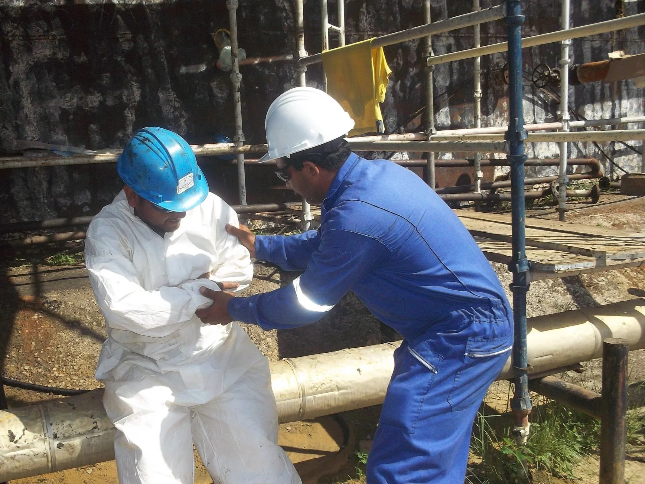Dos trabajadores con cascos de seguridad, uno con traje blanco y otro con traje azul, ayudan a una persona a ponerse una protección o equipo de seguridad en un sitio de construcción o industrial, con andamios y tuberías visibles en el fondo.