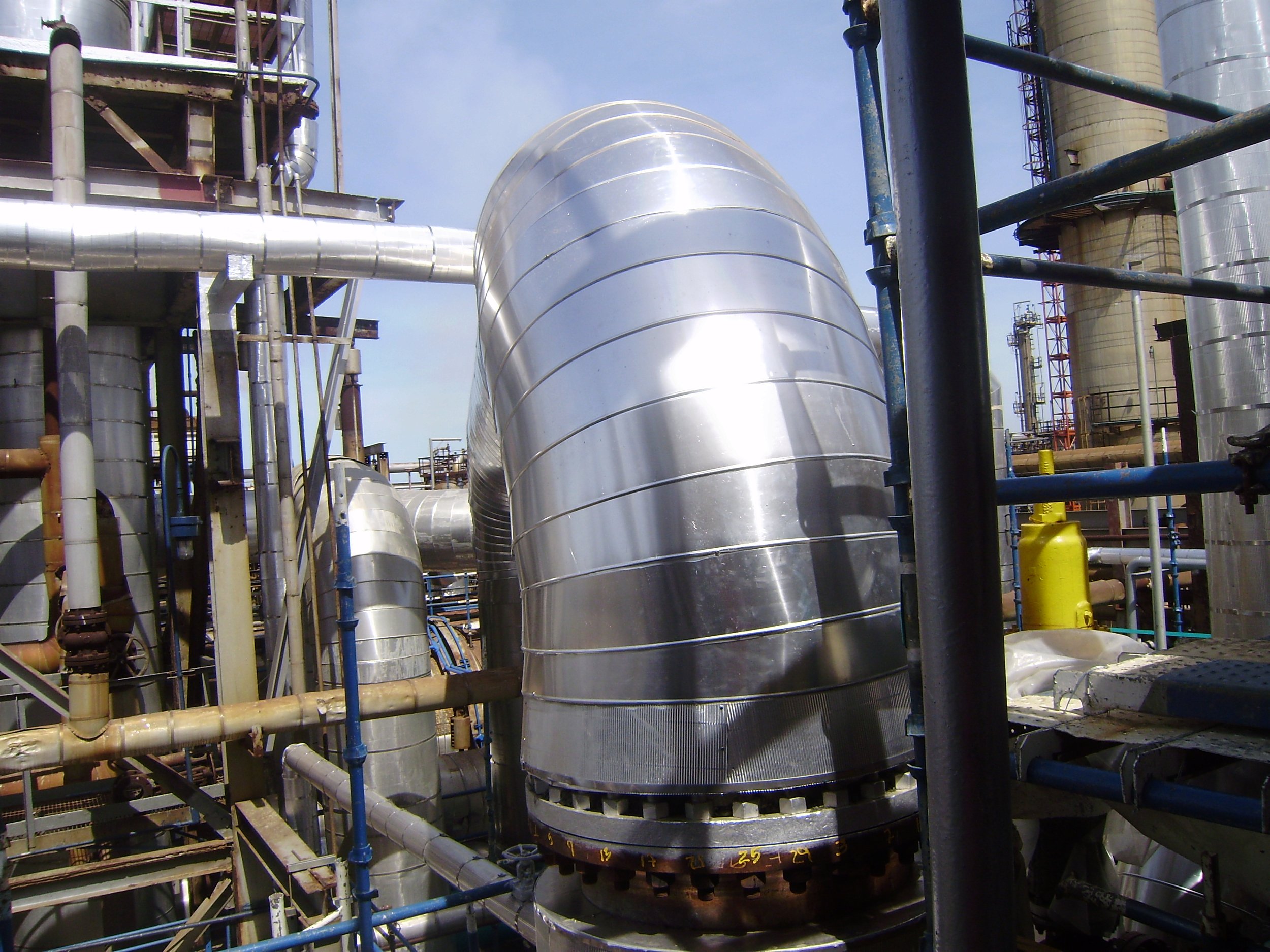 Conjunto de tuberías metálicas en una planta industrial, incluyendo una gran tubería curvada y otras más pequeñas, rodeadas de barandillas y estructuras metálicas.