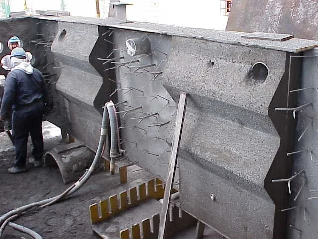 Dos trabajadores en equipo usando casco, gafas y máscara de protección, trabajando en una Estructura de acero con anclajes refractarios en un sitio de construcción.