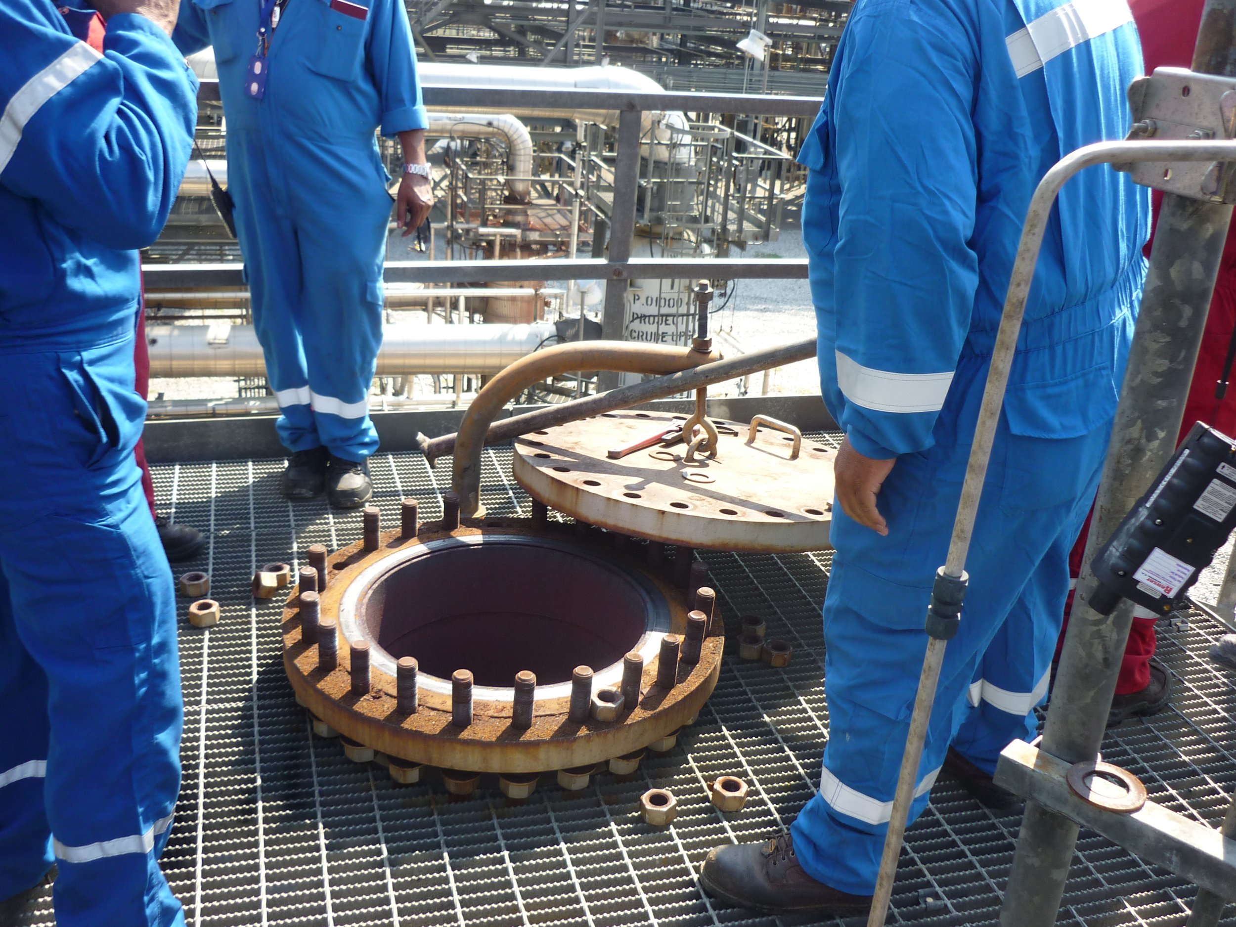Trabajadores de industria en uniforme azul trabajando en una plataforma metálica con maquinaria industrial y tuberías.