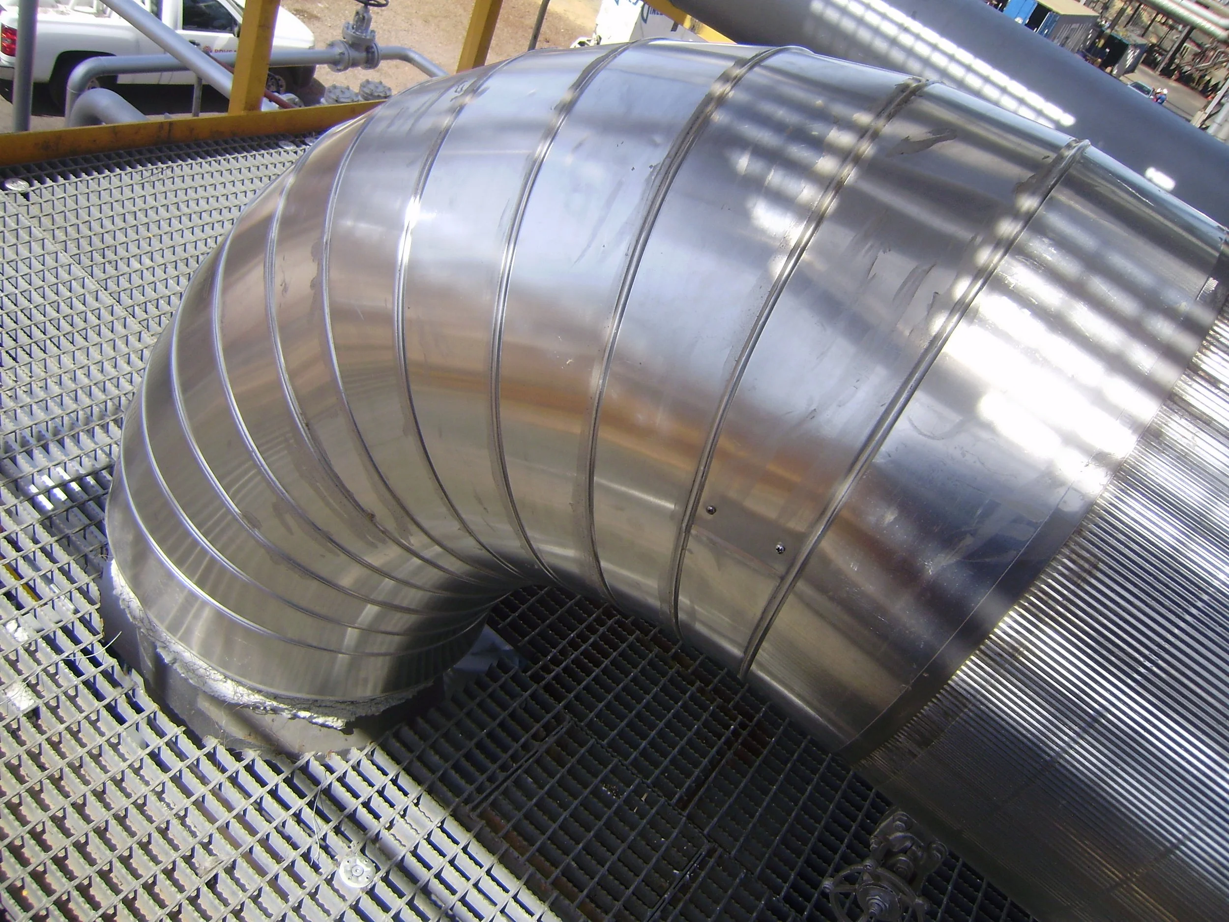 Tubería de acero inoxidable doblada en forma de codo, instalada en una estructura metálica en un entorno industrial.