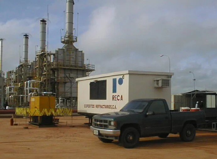 Camióneta verde estacionado frente a una estructura de industria química o petroquímica con chimeneas y maquinaria.