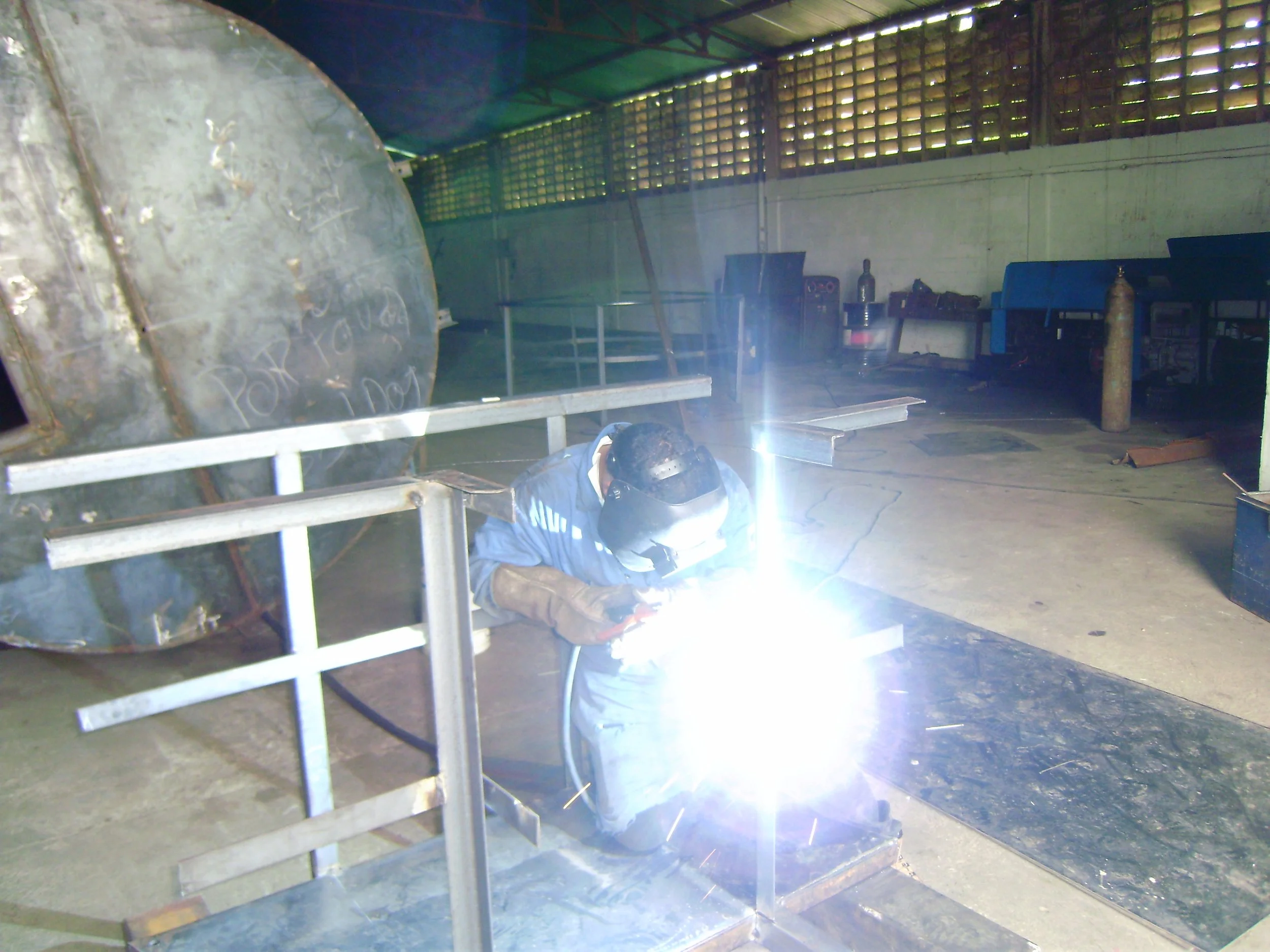 Un trabajador en un taller está soldando una estructura de metal, con protección y arco de soldadura encendido.