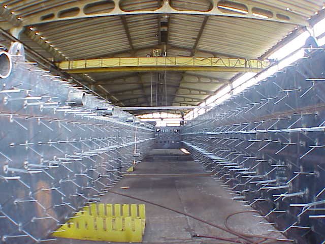 Vista de un interior de un hangar o almacén con estructuras metálicas y un techo de láminas metálicas, con una línea de distanciamiento y algunas herramientas en el suelo.