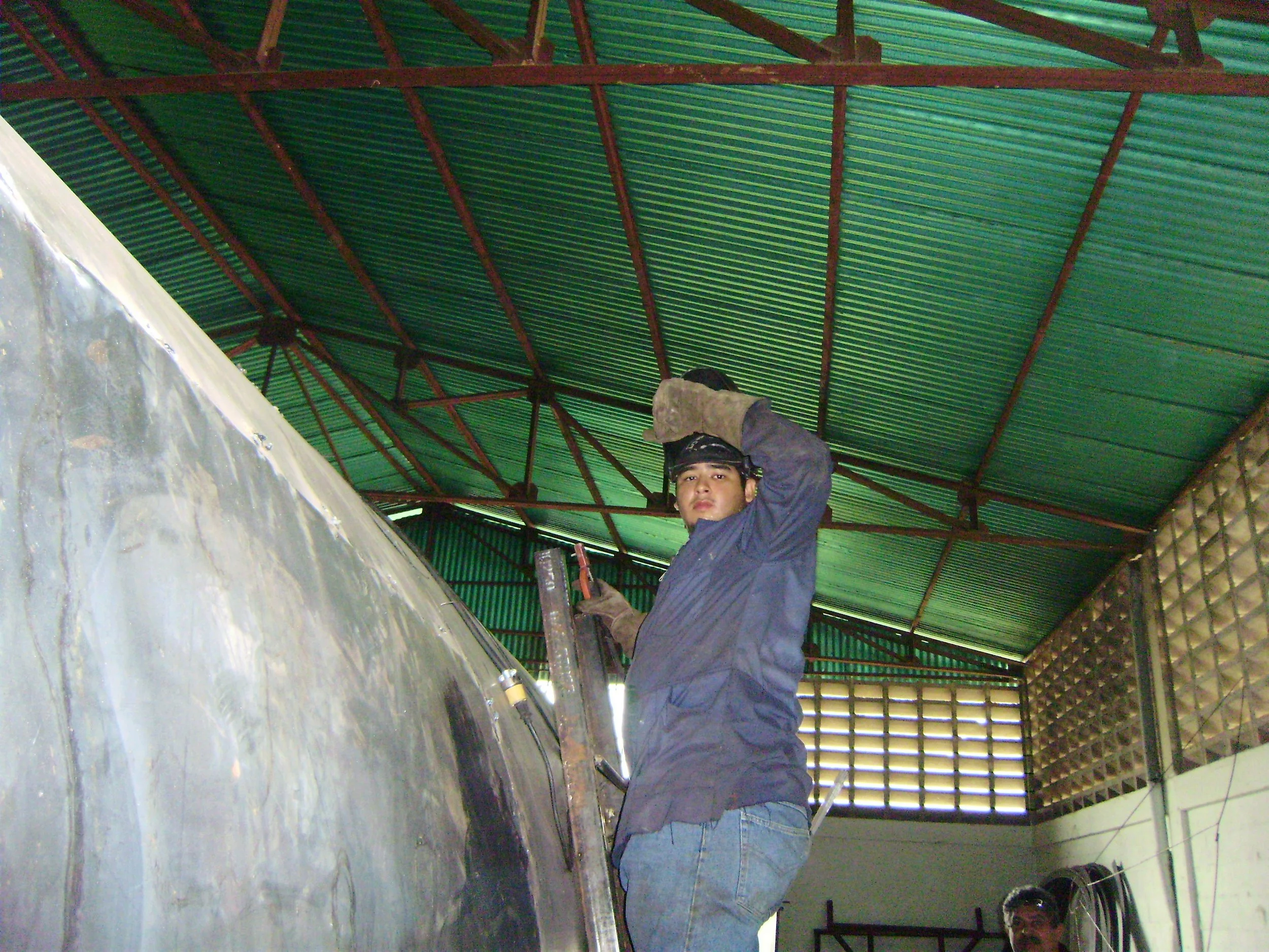Un joven soldador usando guantes y casco de seguridad, trabajando con equipo en un taller con techo de lámina verde.