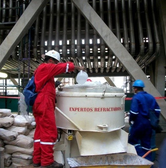 Dos trabajadores en uniformes de protección, uno en rojo y otro en azul, operan en una planta de procesamiento industrial utilizando equipo especializado.