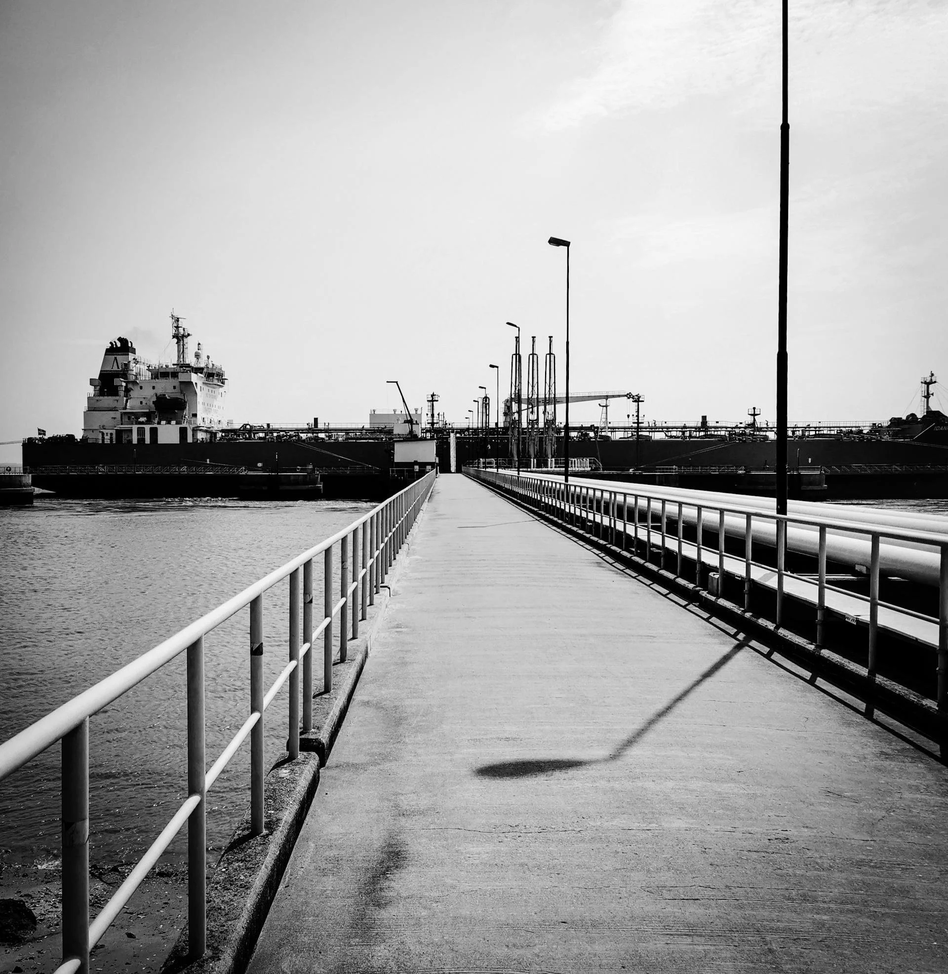 Muelle de embarque con un barco grande en el fondo, postes de luz y tuberías a un lado, en un entorno marítimo en blanco y negro.