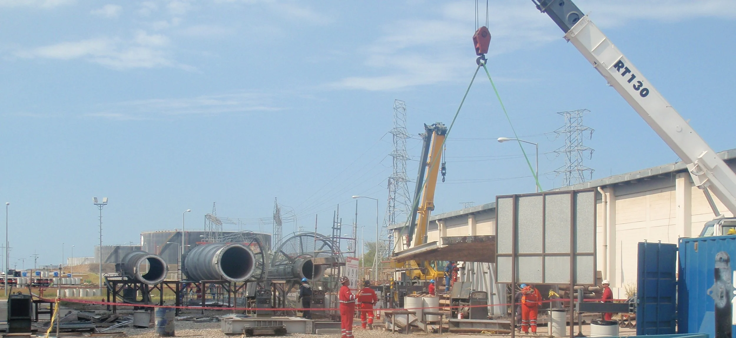 Trabajadores en una construcción en un sitio industrial con equipos pesados, tubos grandes y torres eléctricas en el fondo.